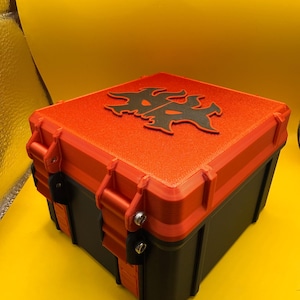 Könnte beinhalten: Eine quadratische Aufbewahrungsbox mit schwarzer Basis und leuchtend orangefarbenem Deckel. Der Deckel zeigt in der Mitte ein erhabenes schwarzes grafisches Emblem. Die Box wird durch orangefarbene seitliche Verschlüsse mit sichtbaren Schrauben gesichert.