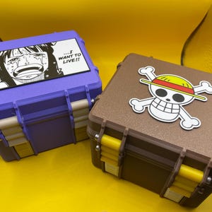 May include: A purple storage case with a black and white graphic of a crying character and the text I WANT TO LIVE, and a brown storage case with a white, black, red, and yellow skull and crossbones emblem. Both cases feature yellow latches.