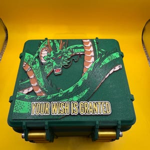 Puede incluir: Caja verde con un diseño de dragón y el texto "YOUR WISH IS GRANTED". El dragón es verde y marrón, y la caja tiene cierres dorados. La caja está sobre un fondo amarillo.