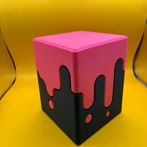 Puede incluir: Un recipiente cúbico con una base negra mate y una tapa rosa brillante. El diseño de la tapa imita un efecto de líquido que gotea, con gotas rosas que se extienden por los lados negros. La superficie del cubo parece lisa y sólida.