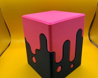 Puede incluir: Un recipiente cúbico con una base negra mate y una tapa rosa brillante. El diseño de la tapa imita un efecto de líquido que gotea, con gotas rosas que se extienden por los lados negros. La superficie del cubo parece lisa y sólida.