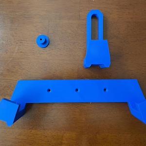 May include: A blue plastic wall mount with three holes and a separate blue plastic hook. The mount is designed to hold a device, such as a phone or tablet.