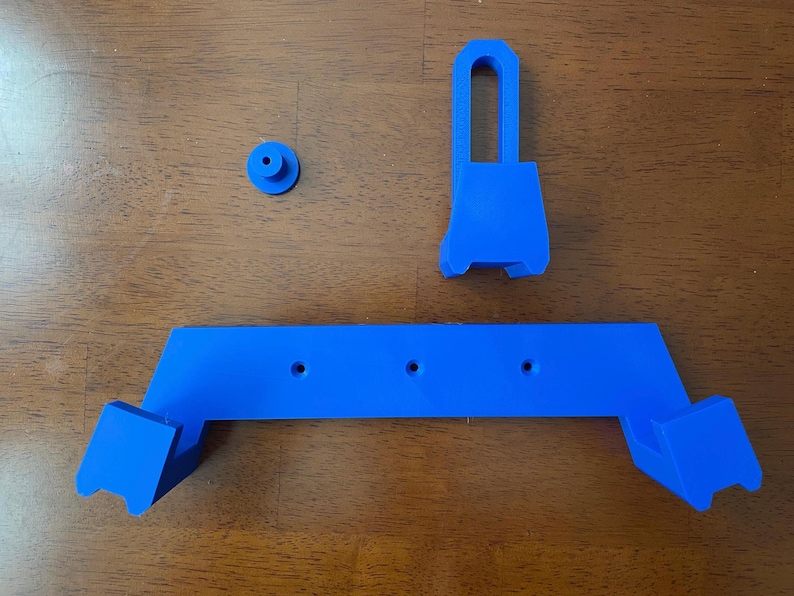 May include: A blue plastic wall mount with three holes and a separate blue plastic hook. The mount is designed to hold a device, such as a phone or tablet.