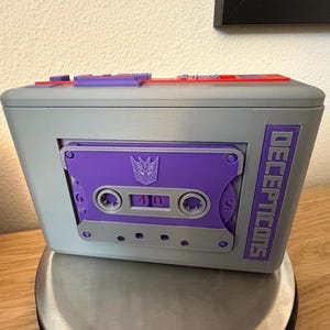 May include: A grey rectangular storage box featuring a purple cassette tape design on the front. The cassette includes the Decepticons logo and the text DECEPTICONS printed vertically. The top of the box has purple and red plastic accents.