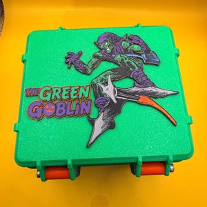 Op de afbeelding: Een groene opbergkoffer met oranje sluitingen en een afbeelding van de Green Goblin. De Green Goblin wordt afgebeeld in een dynamische pose, rijdend op een zweefvliegtuig. De tekst "THE GREEN GOBLIN" is weergegeven in paars en groen.