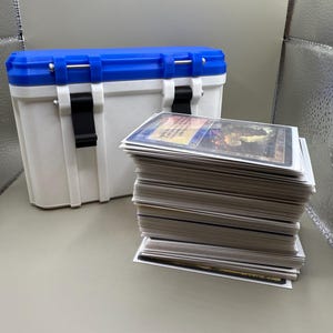 May include: A white and blue storage container with a black latch and a stack of white trading cards.