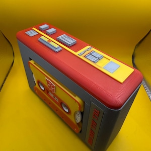 May include: A 3D printed cassette player model in red, yellow, and grey. The top features buttons and a volume slider. A yellow cassette tape with a red Autobot symbol is inserted in the front. The side text reads TILL ALL ARE ONE.