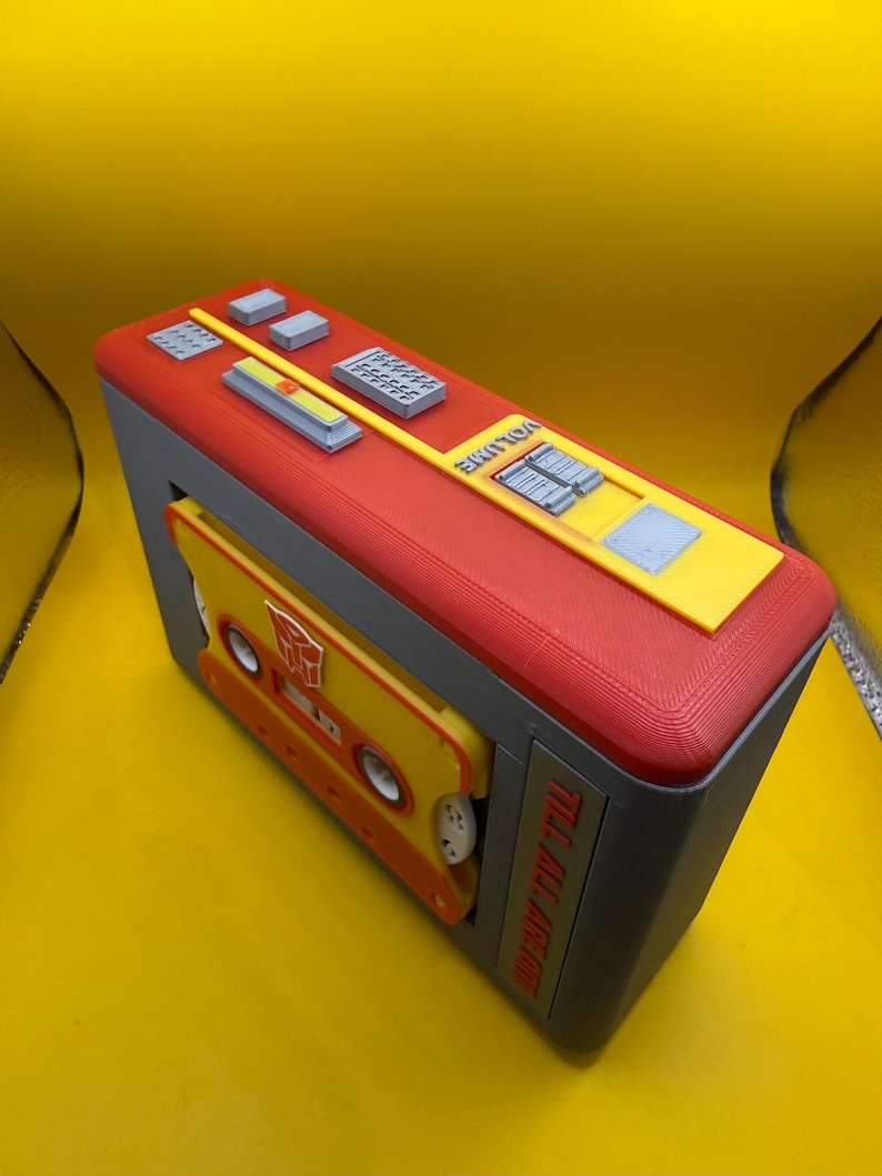 May include: A 3D printed cassette player model in red, yellow, and grey. The top features buttons and a volume slider. A yellow cassette tape with a red Autobot symbol is inserted in the front. The side text reads TILL ALL ARE ONE.