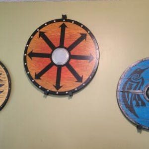 May include: Three round wooden shields with painted designs. The shield on the left has a black raven design on a light brown background. The shield in the center has a black eight-pointed star design on a red and orange background. The shield on the right has a blue and white design with black birds on each side.