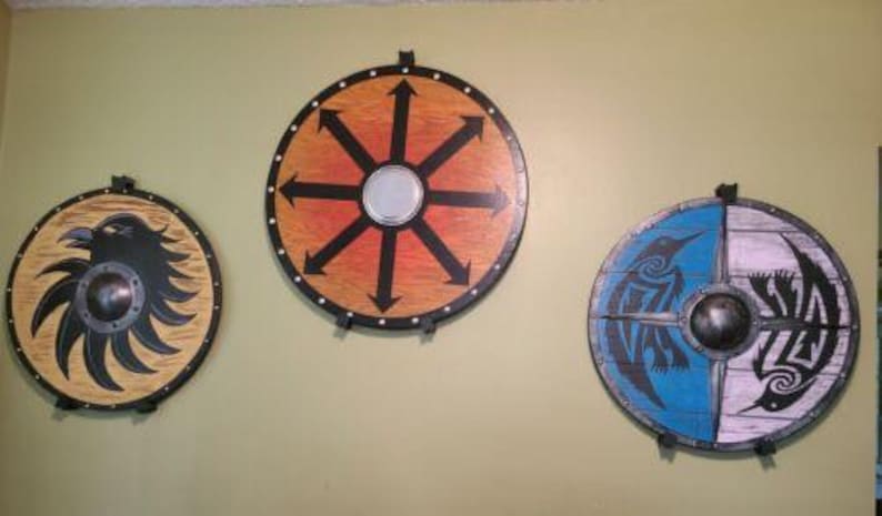 May include: Three round wooden shields with painted designs. The shield on the left has a black raven design on a light brown background. The shield in the center has a black eight-pointed star design on a red and orange background. The shield on the right has a blue and white design with black birds on each side.