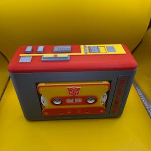 May include: A rectangular red and grey plastic cassette player case featuring a yellow cassette tape insert with a red Autobot logo. The top panel includes grey buttons and a yellow volume slider. The text TILL ALL ARE ONE is printed on the side.