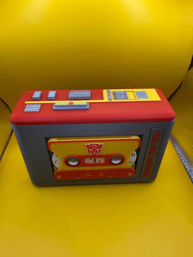 May include: A rectangular red and grey plastic cassette player case featuring a yellow cassette tape insert with a red Autobot logo. The top panel includes grey buttons and a yellow volume slider. The text TILL ALL ARE ONE is printed on the side.