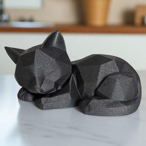 May include: A black geometric cat sculpture in a resting pose. The cat figurine is made of a textured material and has a faceted design. The sculpture is displayed on a white surface.