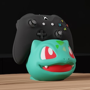 Puede incluir: Un soporte para mando con forma de Bulbasaur turquesa, con ojos rojos y boca sonriente, sosteniendo un mando de Xbox negro. El soporte está sobre una superficie de madera con un fondo oscuro.