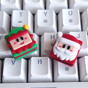 Puede incluir: Dos teclas con temática navideña, una que representa un elfo y otra a Papá Noel, descansan sobre un teclado blanco. La tecla del elfo es verde, roja y beige, mientras que la tecla de Papá Noel es roja, blanca y beige. Las teclas del teclado son blancas.