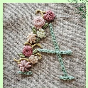 以下が含まれることがあります： Aのアルファベットのクロスステッチ刺繍パターン。ピンク、白、緑の花のフラワーデザイン。パターンはリネン生地の背景です。PDFパターン