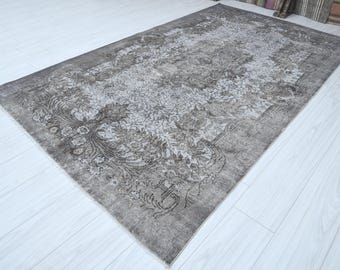 May include: A rectangular, distressed-look rug with a floral pattern in shades of gray and brown. The rug has a faded appearance, with a detailed design featuring stylized flowers and leaves. The rug is placed on a light-colored wooden floor.