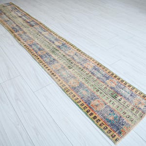 May include: A long, rectangular rug with a faded, vintage look. The rug has a colorful, abstract pattern with shades of blue, green, yellow, and orange. The edges of the rug are trimmed with a dark brown border.