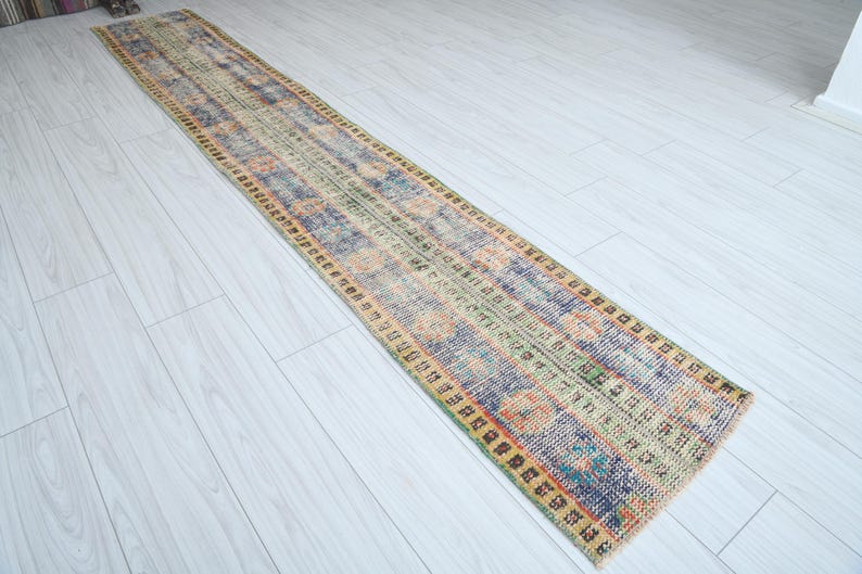 May include: A long, rectangular rug with a faded, vintage look. The rug has a colorful, abstract pattern with shades of blue, green, yellow, and orange. The edges of the rug are trimmed with a dark brown border.