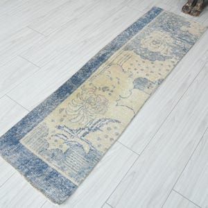 May include: A rectangular, distressed-look rug with a floral design. The rug features a cream-colored central field with blue floral motifs and a blue border. The rug is placed on a light-colored wooden floor.