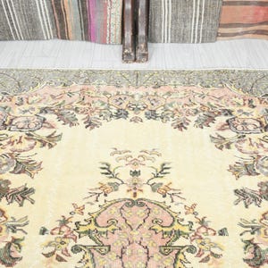 K&ouml;nnte beinhalten: Ein Vintage-Teppich mit cremefarbenem Hintergrund und einem floralen Muster in Rosa-, Gr&uuml;n- und Braunt&ouml;nen. Der Teppich hat einen verblassten, abgenutzten Look, der ihm einen einzigartigen und charmanten Charakter verleiht.