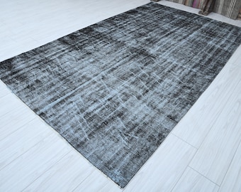 May include: A rectangular, distressed-look rug in shades of black and gray. The rug has a faded appearance with lighter gray streaks running across the surface. The rug is placed on a light-colored wooden floor, with a glimpse of striped fabric in the background.