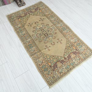 May include: A rectangular beige rug with an intricate central design and border. The rug features a central medallion with floral and geometric patterns in shades of brown, blue, and green. The border has a repeating pattern with similar colors.