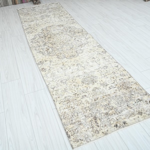 May include: A rectangular, cream-colored runner rug with an intricate, faded floral pattern. The rug's design features a central medallion and detailed borders in shades of brown and gray. The rug is placed on a light-colored wooden floor.