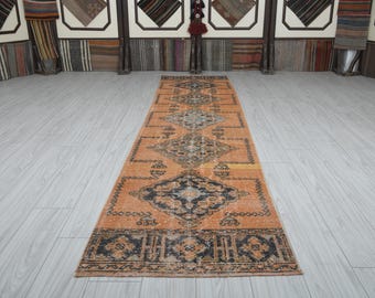 May include: An orange and black patterned runner rug. The rug features a central diamond design with a repeating border pattern. The rug is placed on a light gray wooden floor.