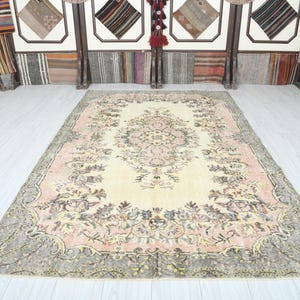 K&ouml;nnte beinhalten: Ein Vintage-Teppich mit cremefarbenem Hintergrund und einem floralen Muster in Rosa-, Braun- und Gr&uuml;nt&ouml;nen. Der Teppich hat einen verblassten, abgenutzten Look, der ihm ein einzigartiges und antikes Aussehen verleiht.