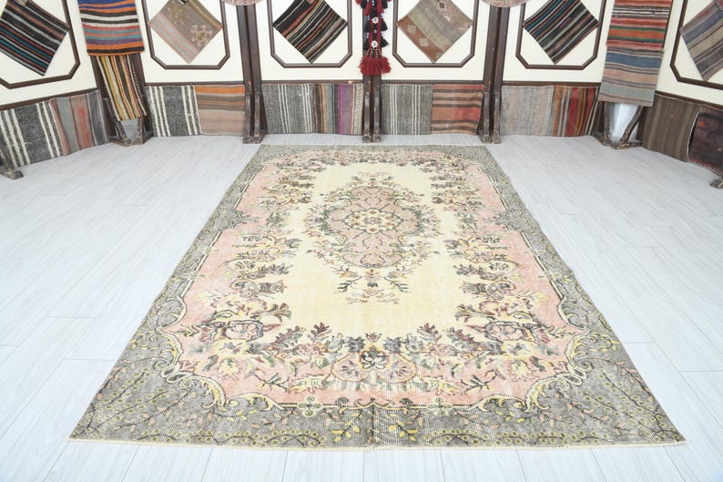 K&ouml;nnte beinhalten: Ein Vintage-Teppich mit cremefarbenem Hintergrund und einem floralen Muster in Rosa-, Braun- und Gr&uuml;nt&ouml;nen. Der Teppich hat einen verblassten, abgenutzten Look, der ihm ein einzigartiges und antikes Aussehen verleiht.
