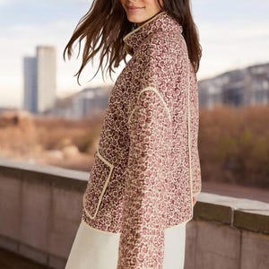 Könnte beinhalten: Burgunderfarbene Jacke mit floralem Muster, hohem Kragen und cremefarbenen Zierleisten. Die Jacke hat eine gesteppte Textur und eine lockere Passform. Das Model trägt einen weißen Rock. Der Hintergrund zeigt eine Stadtlandschaft.