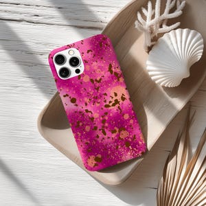 Peut inclure: Coque de téléphone rose avec un motif abstrait vibrant avec des éclaboussures d'or et de marron. L'étui est présenté sur un plateau en bois avec des éléments décoratifs, dont un coquillage blanc et du corail.