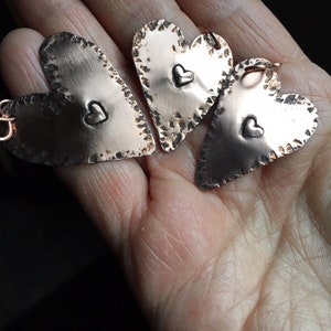 May include: Three copper heart-shaped pendants with a smaller heart design in the center of each. The hearts have a hammered texture and a rustic finish.