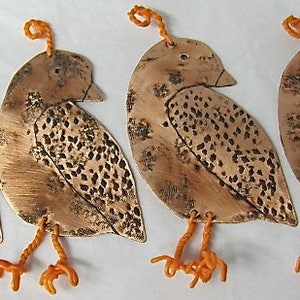 May include: Four copper ornaments shaped like birds with orange string hangers. The birds have a textured surface and are detailed with a pattern of small dots.
