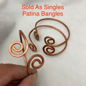 May include: Three copper wire bangles with a spiral design. The bangles are sold individually. The text "Sold As Singles Patina Bangles" is written in white text on a white background.