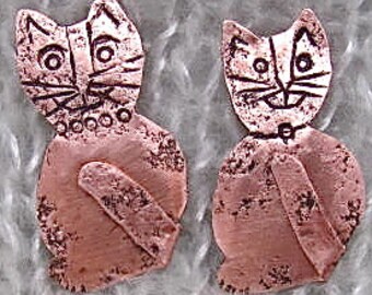 May include: Two copper-colored cat-shaped metal charms. Each charm features a stylized cat design with etched details, including eyes, whiskers, and a collar. The charms have a textured surface and are designed to be used for jewelry making or crafting.