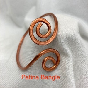 May include: A copper wire bangle with a double spiral design. The bangle has a patina finish. The text "Patina Bangle" is visible below the bangle.