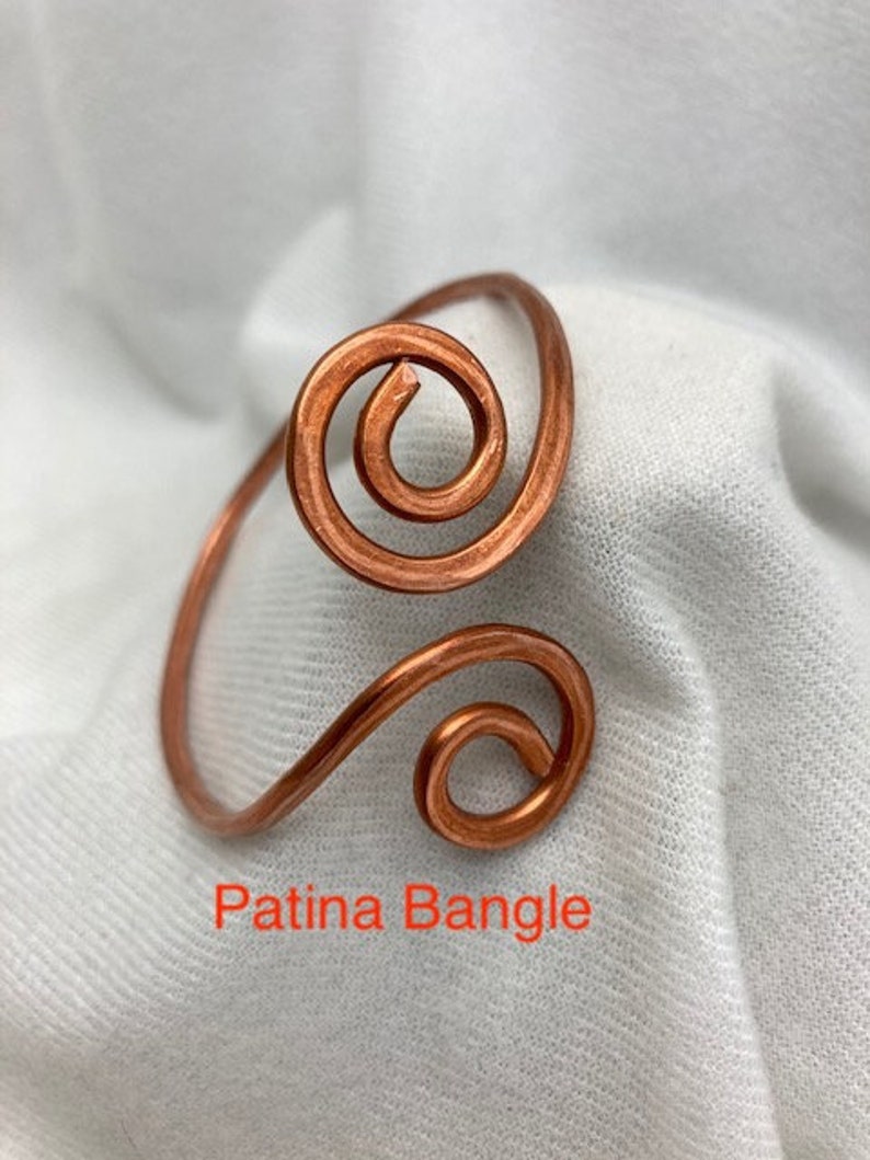 May include: A copper wire bangle with a double spiral design. The bangle has a patina finish. The text "Patina Bangle" is visible below the bangle.