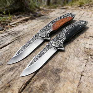 Könnte beinhalten: Zwei Klappmesser mit verzierten silberfarbenen Metallgriffen und Klingen. Ein Messer hat einen Holzgriff mit dem eingravierten Namen "Jenna", und das andere hat einen schwarzen Griff mit den Initialen "KB". Die Messer werden auf einer Holzoberfläche präsentiert.