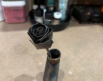 May include: A black metal rose with a curled stem sits in a small, dark blue and gold vase.