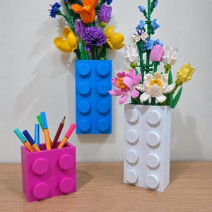 Op de afbeelding: Een felblauwe rechthoekige houder en een witte gespikkelde rechthoekige houder, beide met ronde noppen, bevatten elk een boeket kleurrijke plastic bloemen. Een kleinere roze vierkante houder met noppen bevat diverse gekleurde pennen.