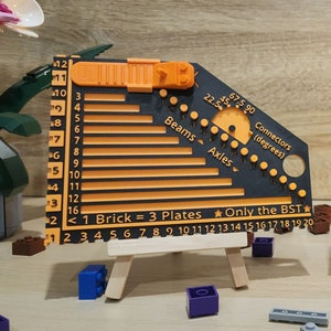 Puede incluir: Dos reglas de plástico negro con marcas naranjas y blancas. Las reglas están etiquetadas como "Beams", "Axles" y "Connectors (degrees)". Las reglas están sobre pequeños caballetes de madera. El texto "1 Brick = 3 Plates" y "Only the BST" está impreso en las reglas.