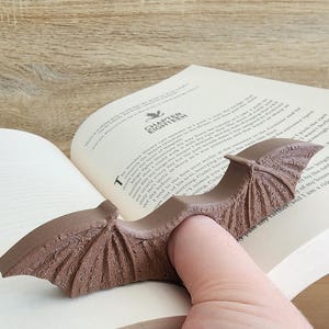May include: A brown, detailed, bat-shaped bookmark with a textured surface. The bookmark is designed to hold a page open with a finger.