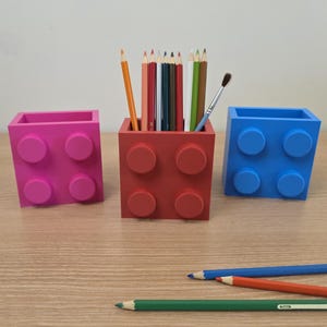 以下が含まれることがあります： ピンク、赤、青のブロック型のカラフルなペン立てが3つ。それぞれに4つの丸いスタッドが付いています。赤いホルダーには鉛筆と筆が入っています。青、オレンジ、緑の鉛筆が木製の表面に散らばっています。