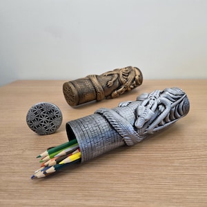 May include: Two decorative pencil holders, one silver and one brown, with dragon and rope designs. The silver holder contains colored pencils. A small, round, patterned object and a pink eraser are also present. The items are on a wooden surface.
