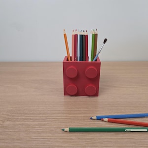 Peut inclure: Un porte-crayons rouge en forme de brique, rempli de crayons de couleur et d'un pinceau. Trois crayons bleus, rouges et verts sont pos&eacute;s sur la surface en bois devant le support. Les crayons sont de couleurs vari&eacute;es.