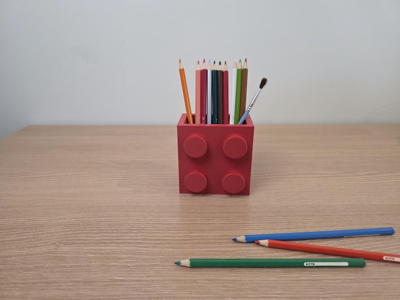 Peut inclure: Un porte-crayons rouge en forme de brique, rempli de crayons de couleur et d'un pinceau. Trois crayons bleus, rouges et verts sont pos&eacute;s sur la surface en bois devant le support. Les crayons sont de couleurs vari&eacute;es.