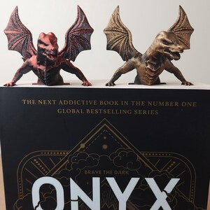 May include: Two dragon bookends in bronze and red hues sit atop a book titled "ONYX STORM". The bookends are detailed with wings and a fierce expression. The book cover has gold lettering and the text "BRAVE THE DARK".