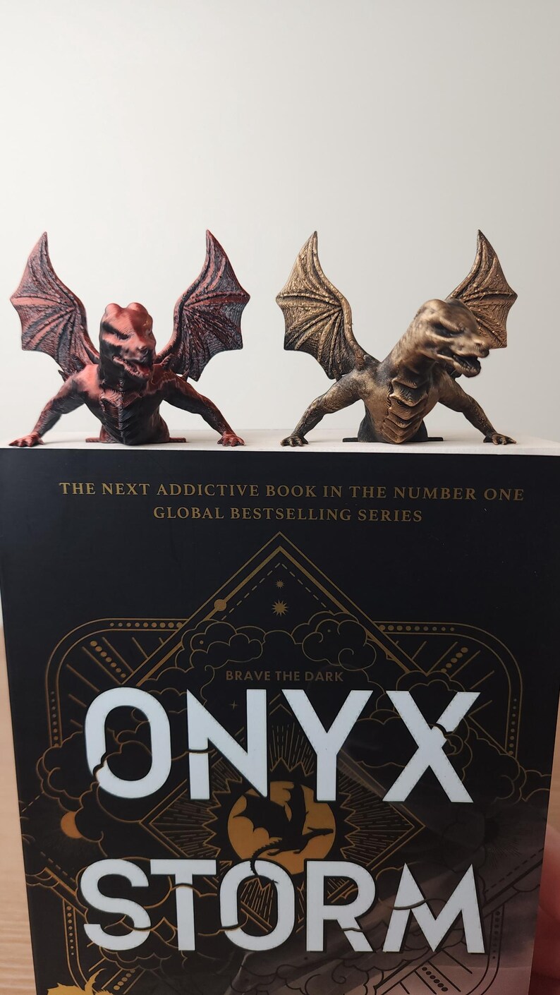 May include: Two dragon bookends in bronze and red hues sit atop a book titled "ONYX STORM". The bookends are detailed with wings and a fierce expression. The book cover has gold lettering and the text "BRAVE THE DARK".